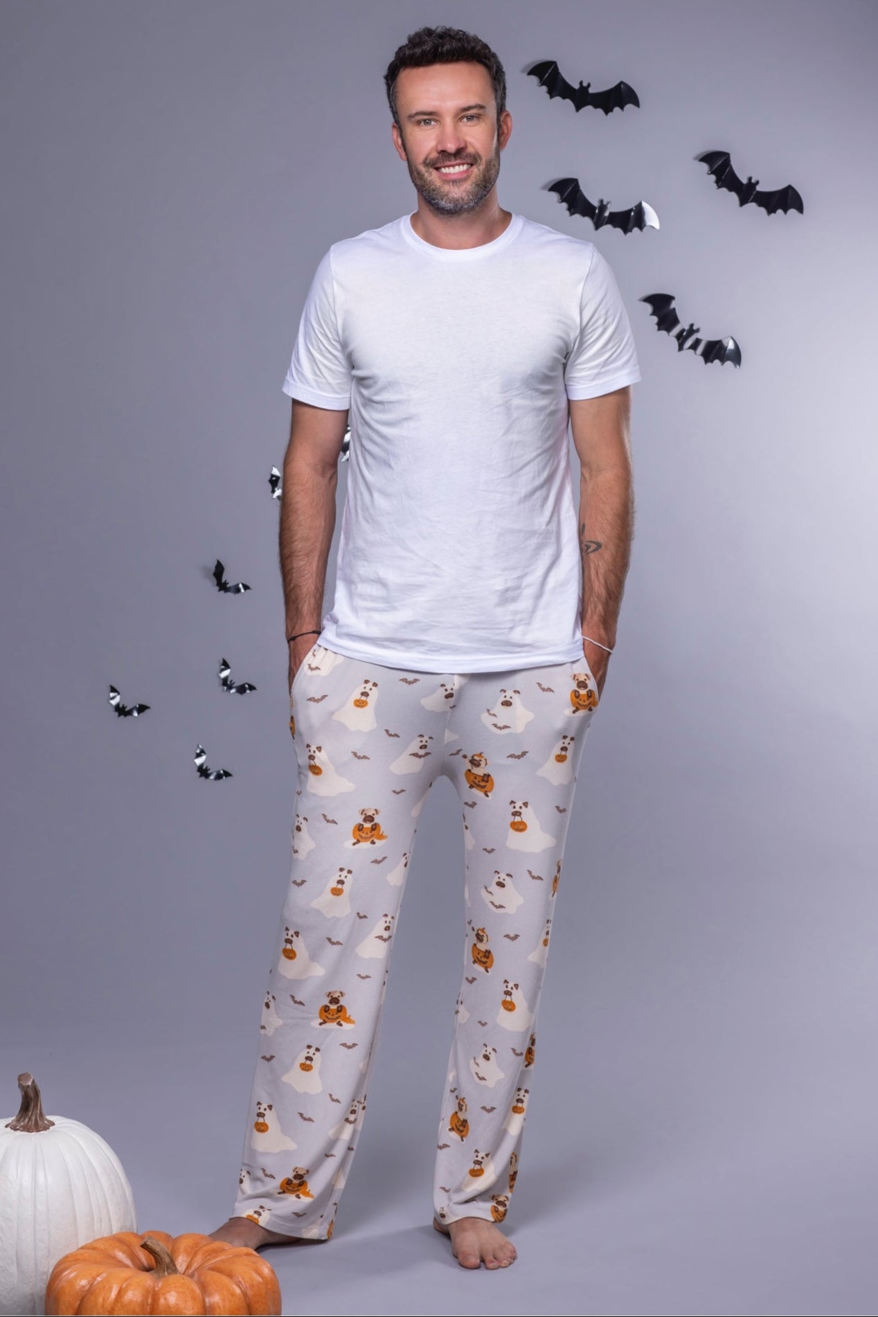 Man wearing a white t-shirt and patterned pajama pants with pumpkins and ghosts, standing against a gray background with decorative elements.